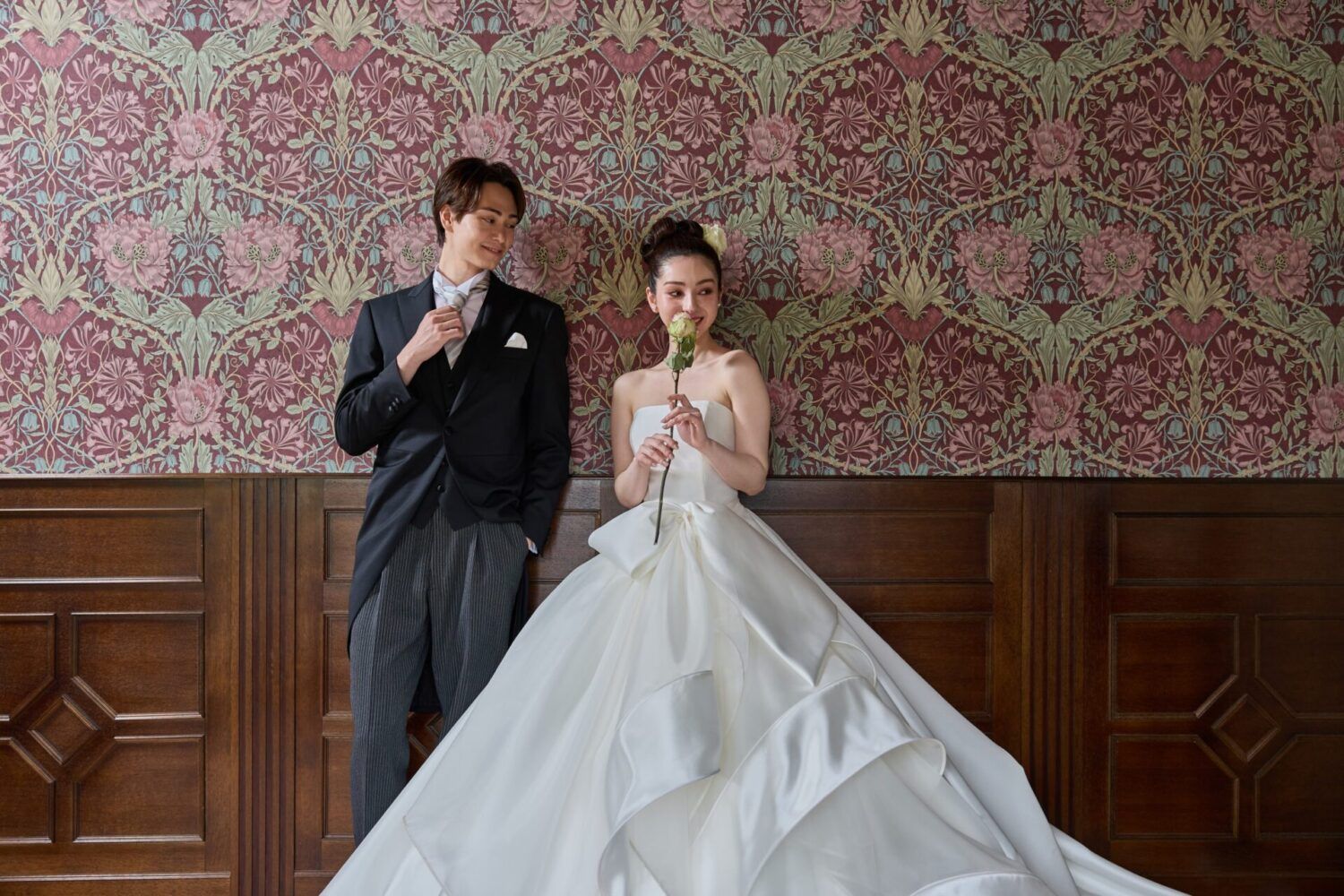 英国アンティークスタジオでウイリアムノリスの壁紙を背景にカンタベッラドレス(Cannta Bella Dress）の花嫁と新郎