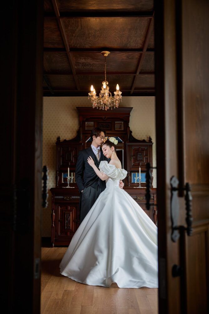 英国アンティークスタジオの燭台を背景に扉越しのカンタベッラドレス(Cannta Bella Dress）の花嫁と新郎
