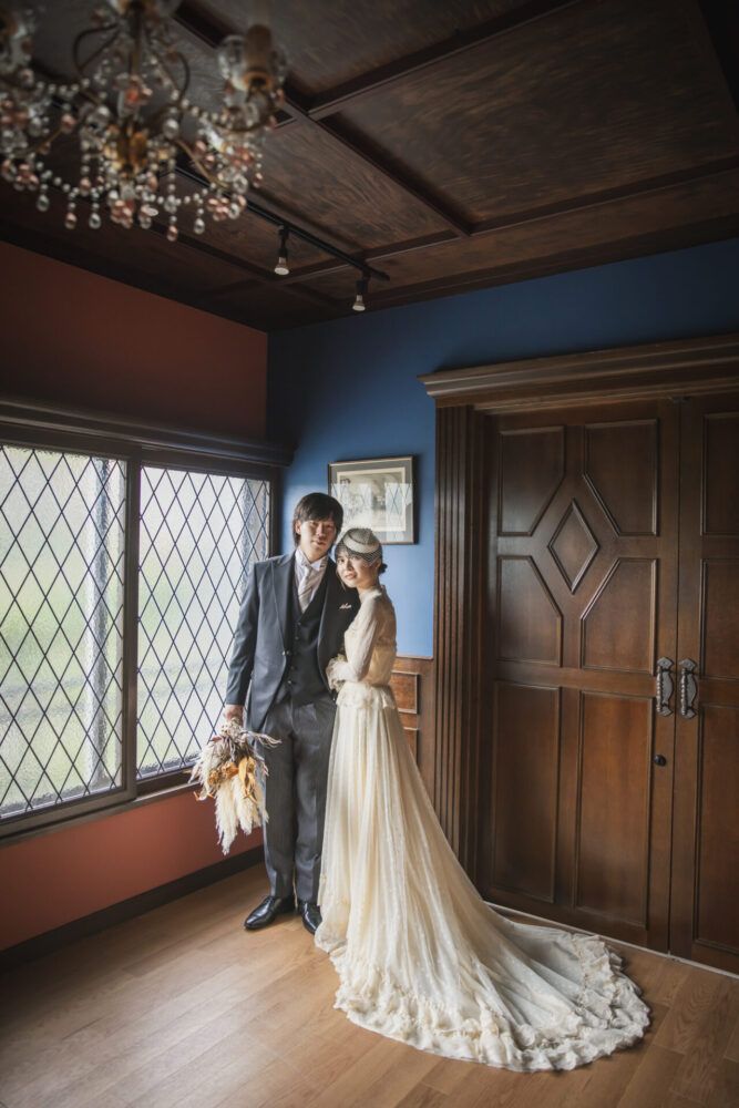 英国アンティークスタジオでステンドグラスと重厚な扉を背景にヴィンテージドレスの花嫁と新郎