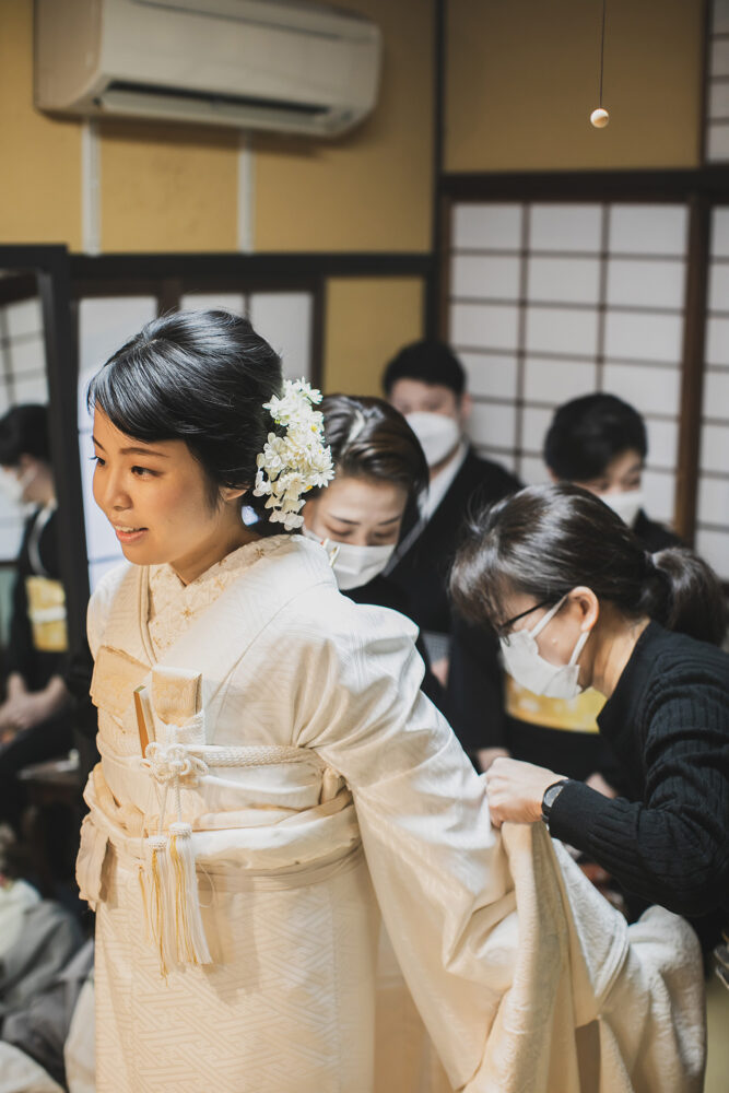 ウエディングフォト:挙式を控え、スタッフに囲まれながら白無垢の着付けを整えてもらう新婦の緊張感と喜びに満ちた表情です。
