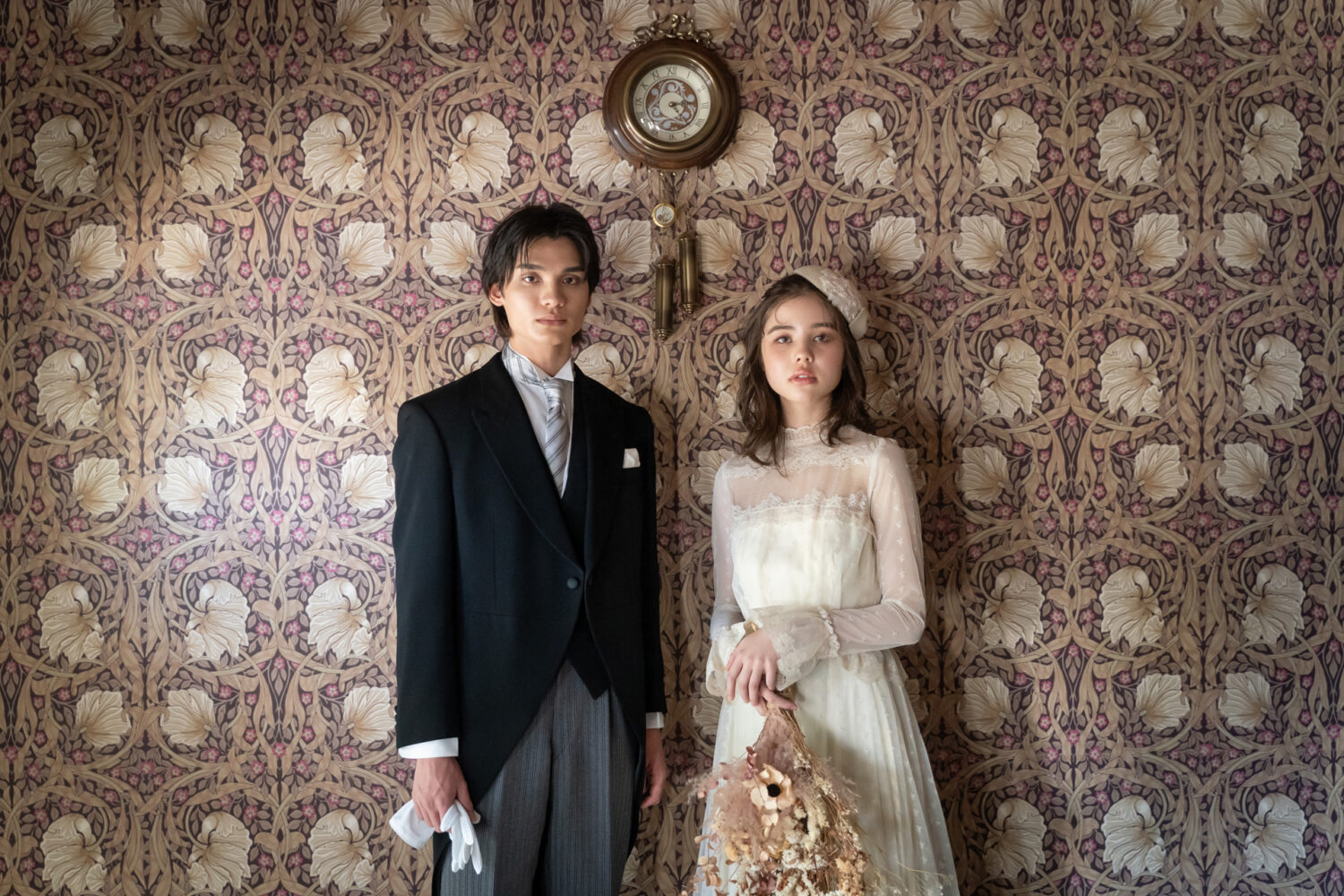 ウィリアム・モリス風の植物柄の壁紙とアンティーク時計を背景に、繊細なレースのヴィンテージドレスとタキシードで並ぶ新郎新婦の大正ロマンなウェディングフォト