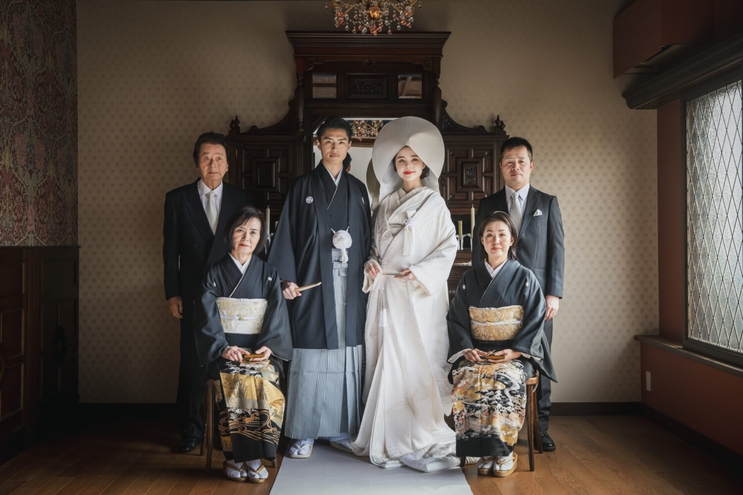 クラシックな洋館スタジオでの家族の集合写真。中央に紋付袴の新郎と白無垢・綿帽子の新婦、両脇に黒留袖の母と黒スーツの父が並んで直立している。