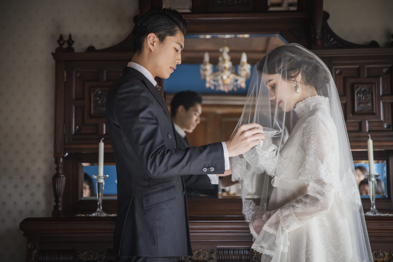 アンティーク家具を背景に、繊細なレースのヴィンテージドレスとタキシードで向かい合う新郎新婦のウェディングフォト