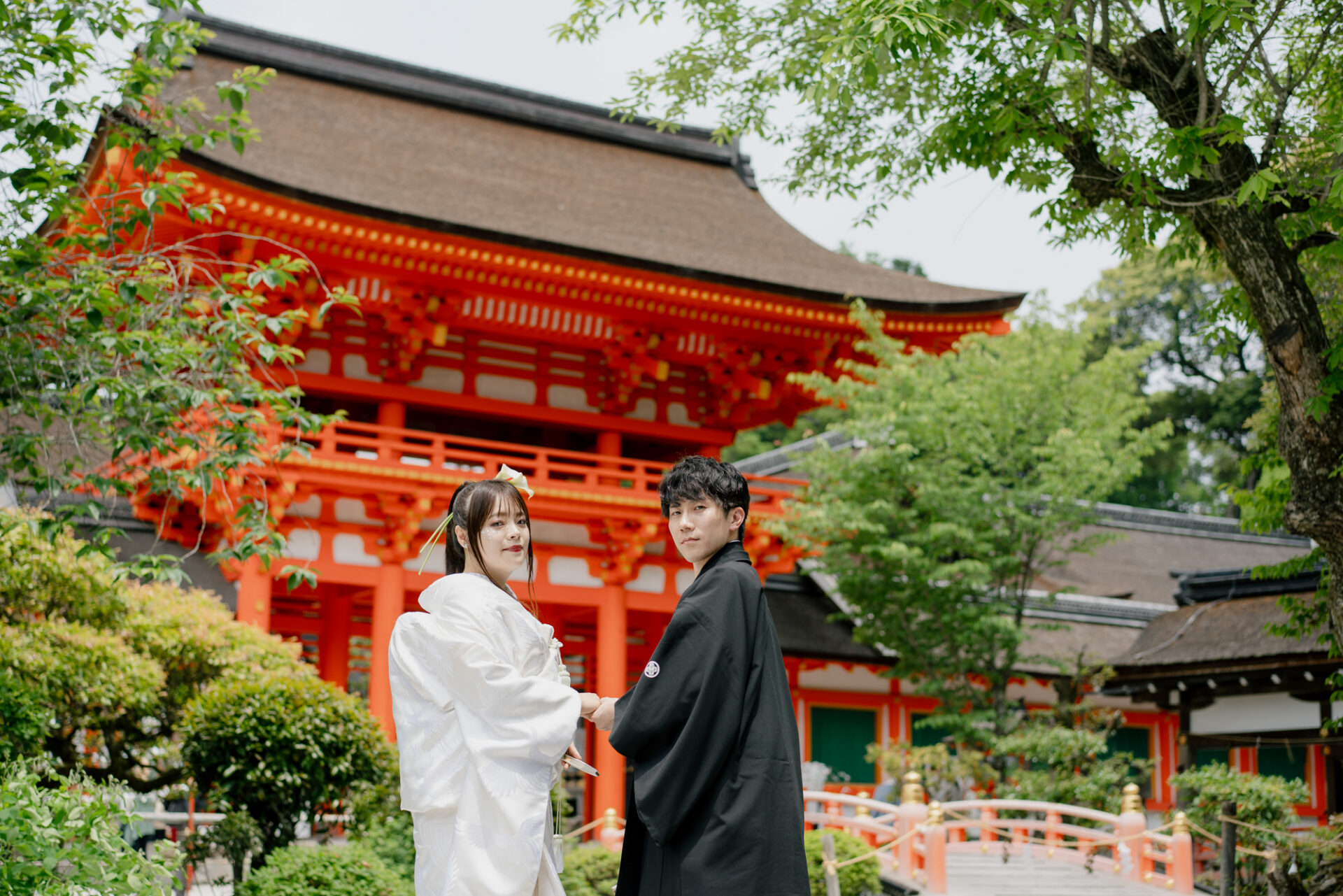 厳かさと華やかさが交差する、上賀茂神社・楼門前でのフォトウェディング
京都最古の神社のひとつ「上賀茂神社」。澄んだ空気と凛とした静けさに包まれるこの場所で、フォトウェディングの一枚一枚が、特別な物語として刻まれていきます。

この日、新郎新婦のお二人が立たれたのは、国の重要文化財にも指定されている**楼門（ろうもん）**前。鮮やかな朱色と白木のコントラストが印象的なこの門は、上賀茂神社を象徴する存在でもあり、写真に残るその姿は、まるで絵巻物のような美しさです。

白無垢姿の新婦様と、紋付袴の新郎様。歴史ある楼門を背景に並ばれるその立ち姿は、まさに「和の美」の極み。新婦様の綿帽子がやわらかな日差しを受けてふんわりと光り、伝統と格式を感じさせるショットに仕上がりました。

楼門前は光の入り方も絶妙で、晴れの日には朱塗りの柱がより一層映え、曇りの日にはしっとりと落ち着いた雰囲気に。どちらの表情も素敵で、撮影するたびに新しい発見があります。

お二人の幸せな門出を、千年の時を越えて見守り続けるこの楼門。そんな特別な場所での一枚は、きっと一生の宝物になることでしょう。

