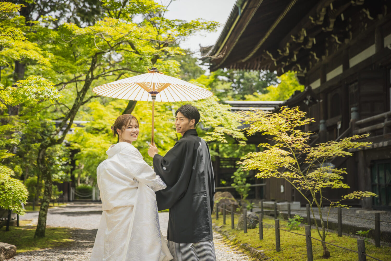 新緑が美しい京都の寺院を背景に、番傘を差し、白い和装と黒の紋付袴姿で見つめ合う新郎新婦のロケーションフォト。