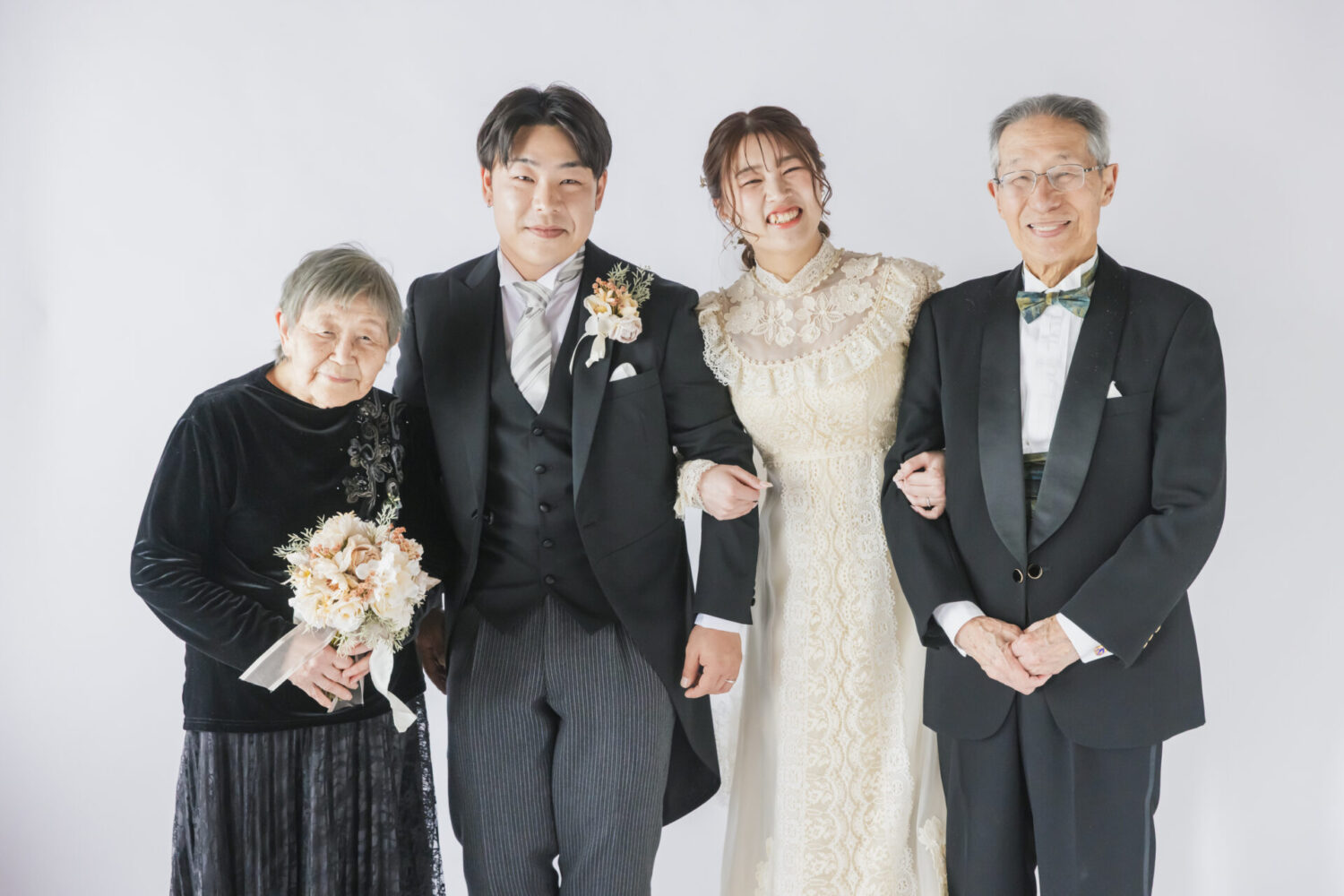 洋装でのフォトウェディング。ドレス姿の新婦とタキシードの新郎が、大切なお祖父母様と腕を組んで笑顔を見せる親孝行カット。