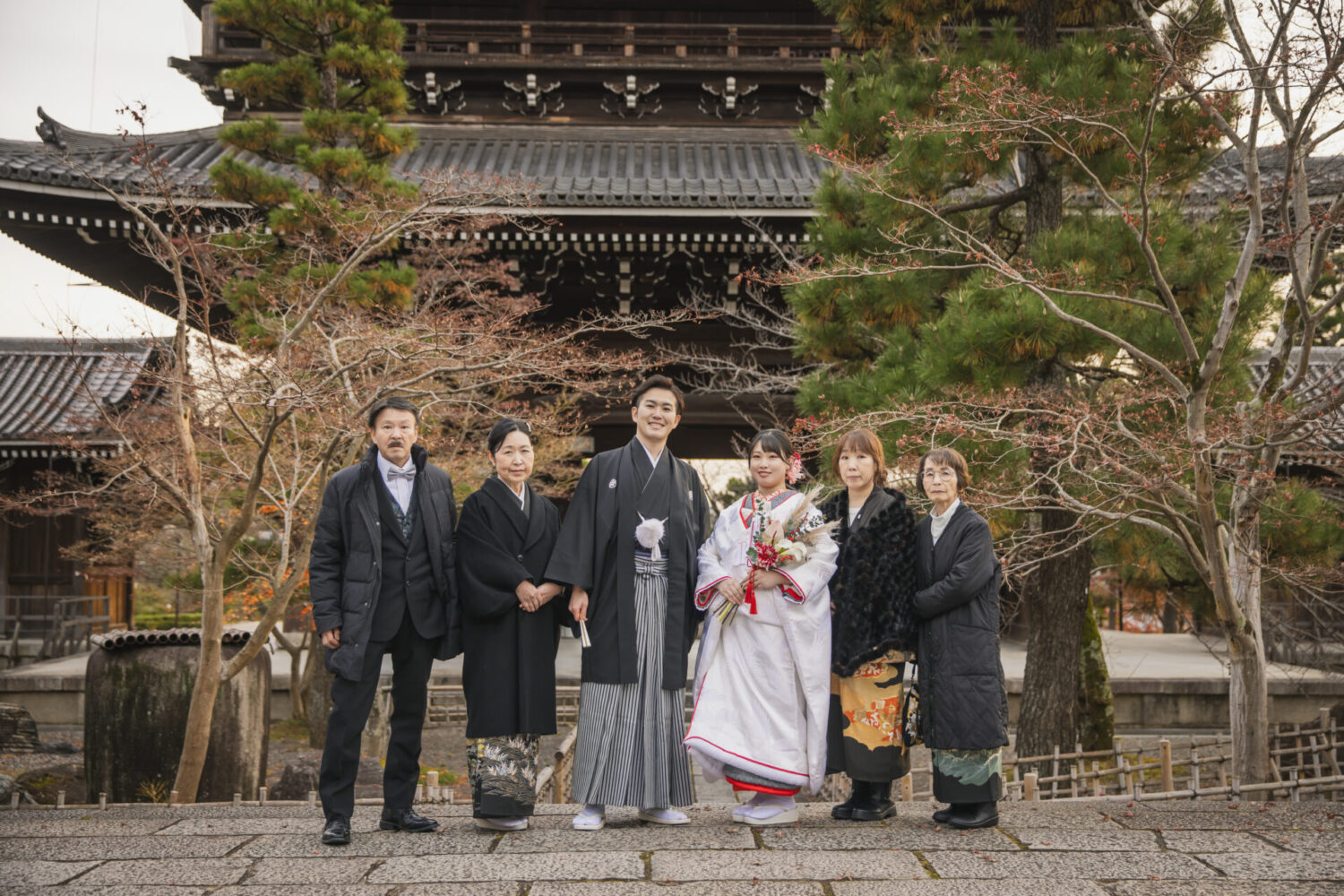京都の歴史ある寺院の門前で、白無垢姿の新婦と紋付袴の新郎、そして黒留袖や正装に身を包んだご家族全員での厳かな集合写真。(家族ウエディングフォト)
