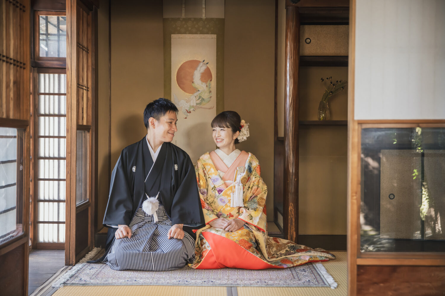 趣のある京町家の畳の間で、色打掛と紋付袴を着用して正座し、和やかな表情で向き合う新郎新婦。