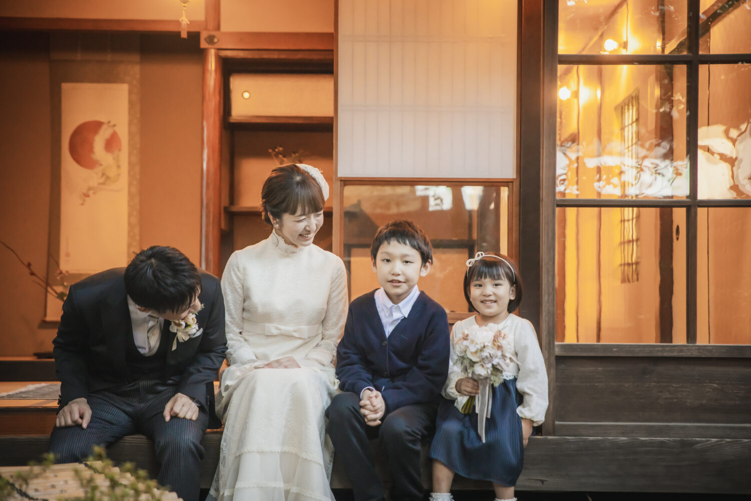 京都町家寫眞館の京町家スタジオの縁側に座る、洋装の新郎新婦と二人の子供。新婦はアンティークな白ドレス、子供は紺色のカーディガンやボレロを着用し、娘が小さなブーケを持っている。
