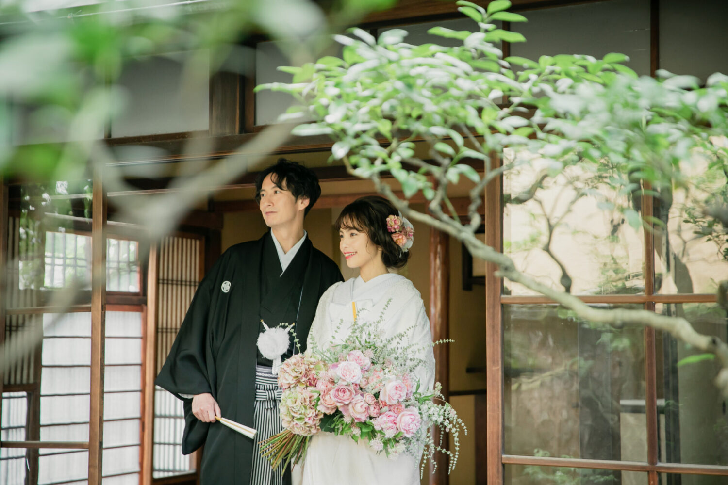 古民家の縁側付近で、大きなピンクのブーケを持った新婦と紋付袴の新郎が寄り添い、庭を眺めている和装ウェディングフォト。