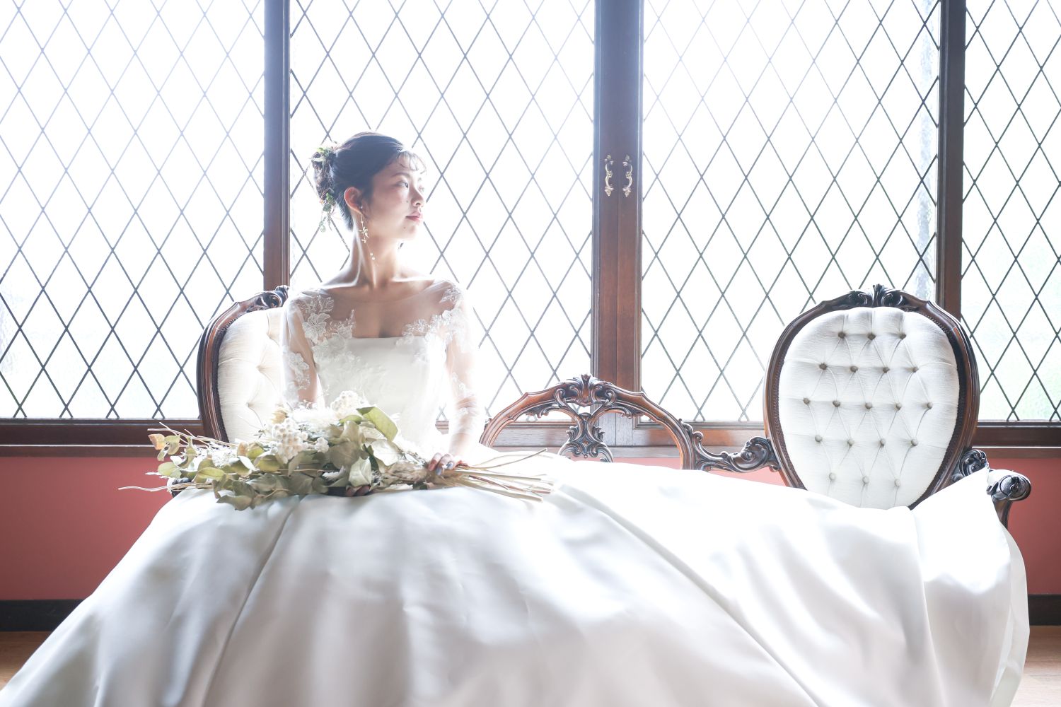 格子窓の光を背に、レースの袖とボリュームのあるスカートのドレスでアンティークの椅子に座る花嫁