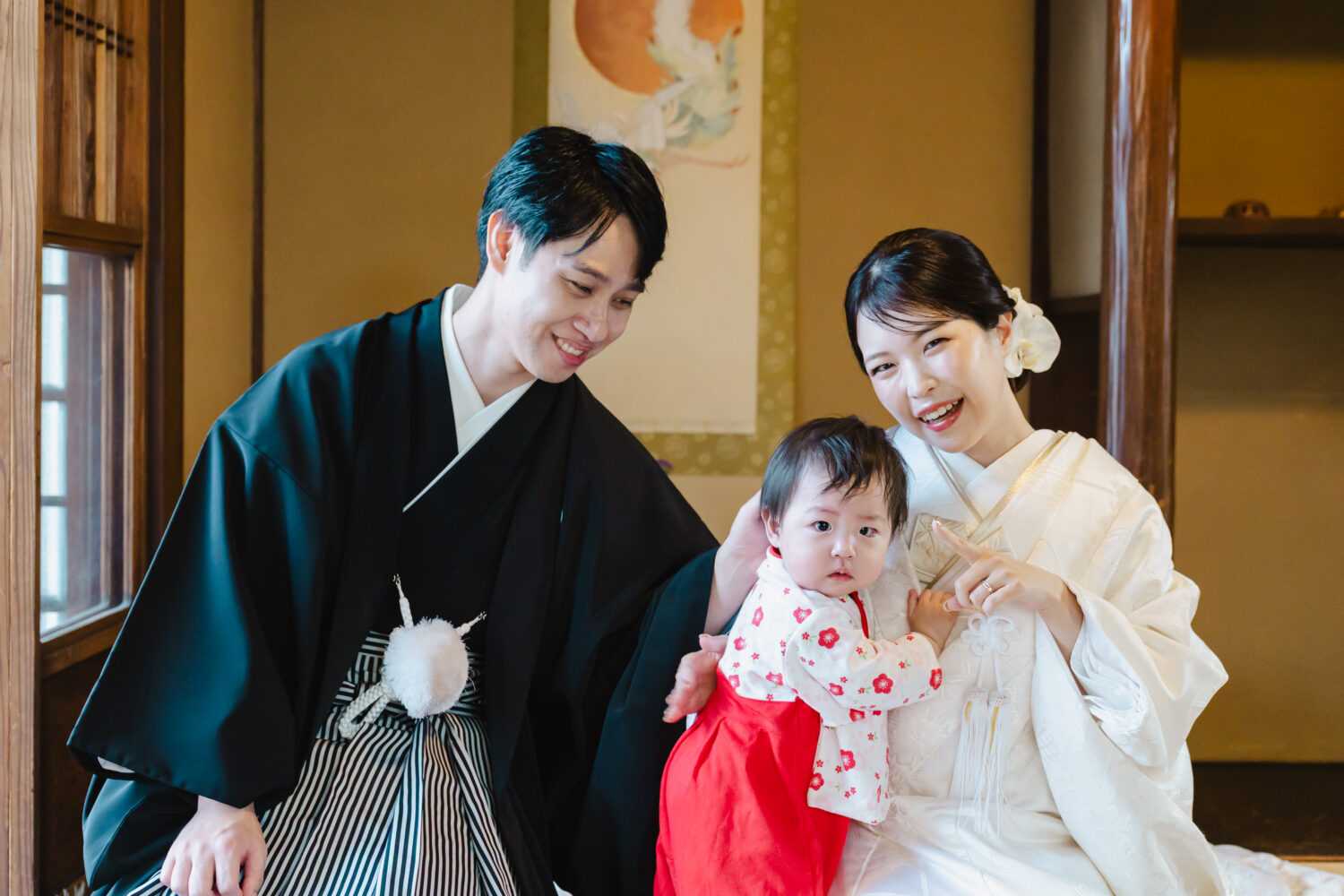 ウエディングフォト:和室にて、紋付姿の新郎と白無垢の新婦が、赤い和装を着た小さなお子様を優しく見守る家族のポートレートです。