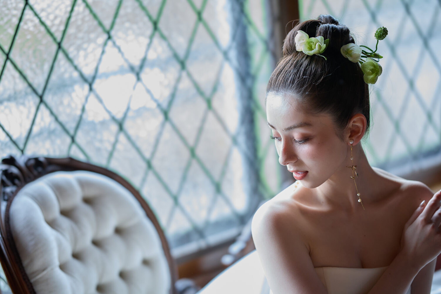 格子窓からの光のなか、生花を挿したアップスタイルに揺れるイヤリングを合わせた花嫁が伏し目で座る