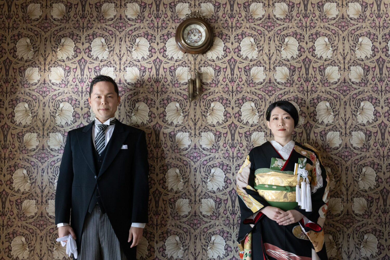 アンティークな壁紙の前で正装して並ぶ、黒引き振袖とタキシードの和洋折衷スタイル