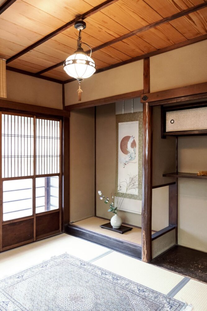 京町家の趣ある和室。 鶴が描かれた掛け軸と季節の花が生けられた床の間、天井から吊るされたアンティークな照明がノスタルジックな空間。