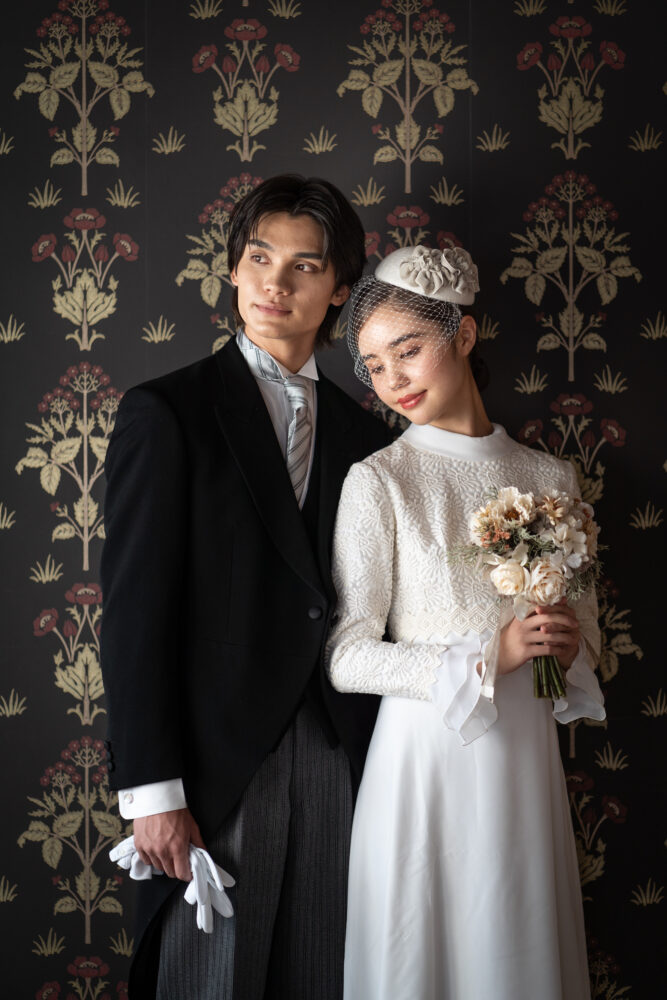 ウィリアム・モリス風のレトロな壁紙を背景に、レースのヴィンテージドレスとトーク帽を合わせた花嫁と、タキシード姿の新郎が佇む大正ロマンなウェディングフォト