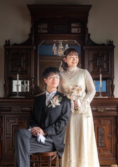 大きなアンティークの鏡台の前に佇む新郎新婦。 温かみのある光の中で、繊細なレースを重ねたハイネックドレスと正装の新郎が優しく微笑むレトロな雰囲気。