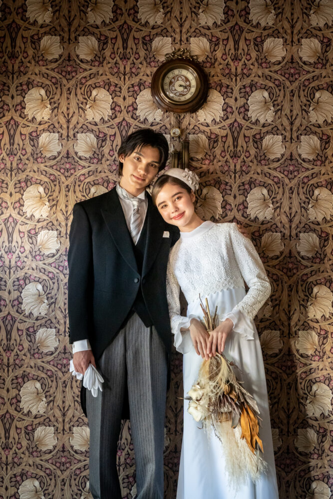 ウィリアム・モリスの壁紙を背景に寄り添う新郎新婦。 新婦はハイネックの総レースドレスにトーク帽、新郎はクラシックなモーニングコートを着用したヴィンテージスタイル。