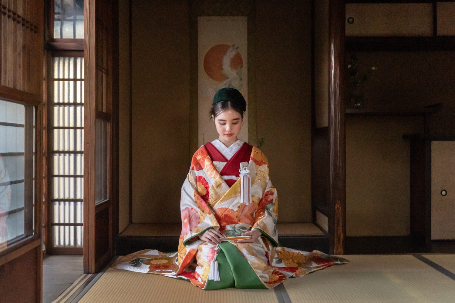 柔らかな光が差し込む和室の床の間で、静かに正座をする新婦のソロショット。 金糸が輝く豪華な色打掛に、深い緑の帽子を合わせたクラシックかつ独創的な和の装い。