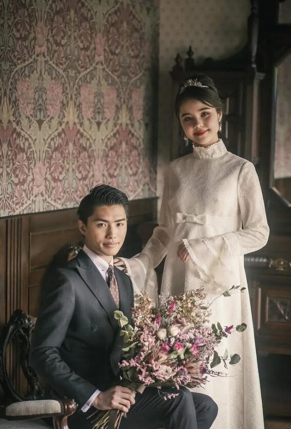 重厚なアンティーク家具を背にした新郎新婦のポートレート。 新婦はパフスリーブのクラシカルな白ドレスにティアラ、新郎はダークスーツを纏い、ドライフラワーのブーケを抱えた一枚。