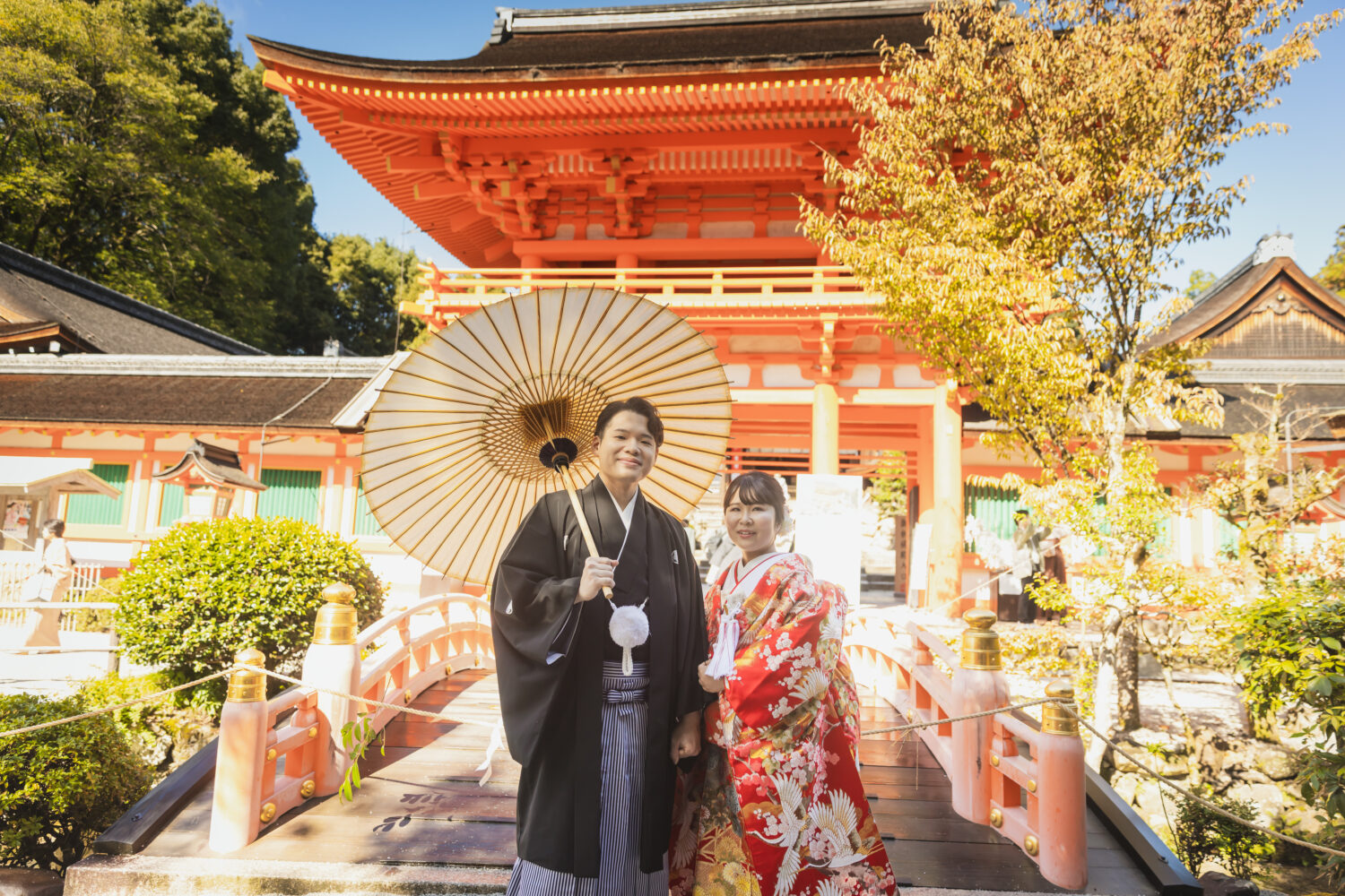 鮮やかな朱色の大きな門と太鼓橋がある神社を背景に、華やかな赤の色打掛と黒の紋付袴で寄り添い立つ新郎新婦。