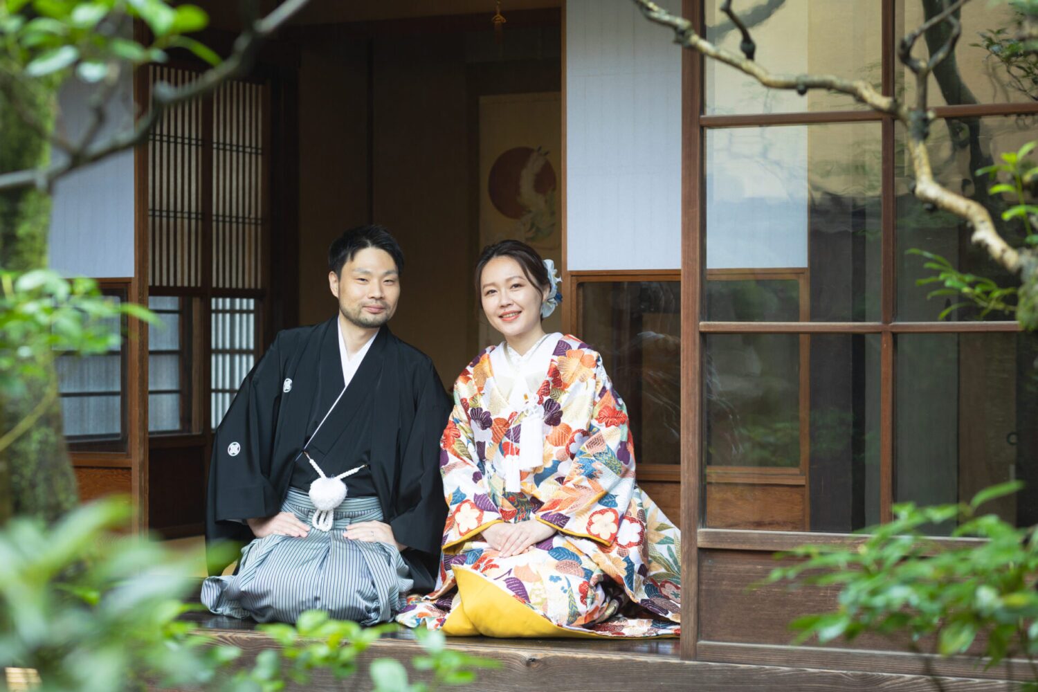 京町家の縁側に並んで座る、和装の新郎新婦。引き振袖姿の新婦と黒紋付の新郎が庭を背景に微笑む
