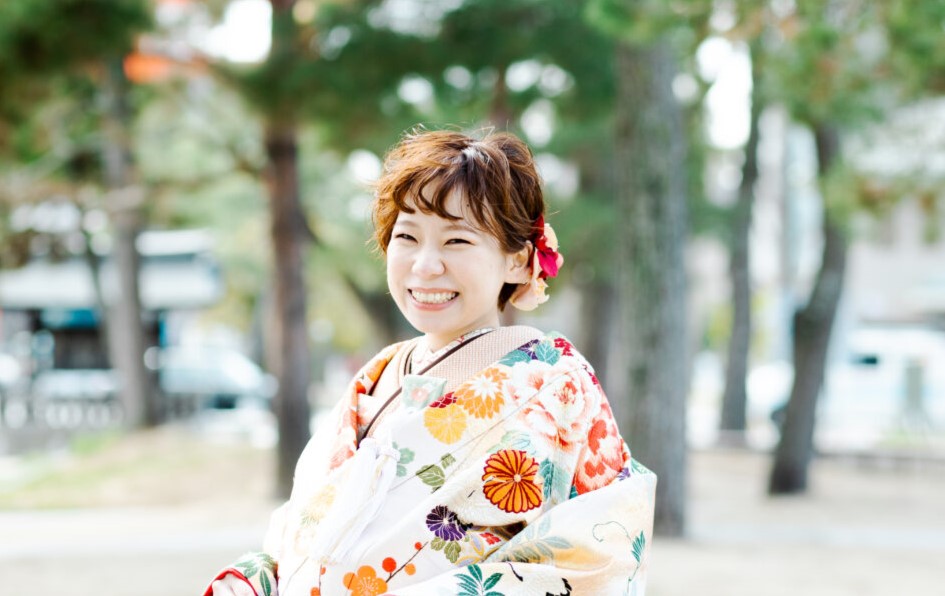 華やかな色打掛を着て、屋外で満面の笑みを浮かべるショートヘアの花嫁。耳元には大きな赤い花の髪飾り