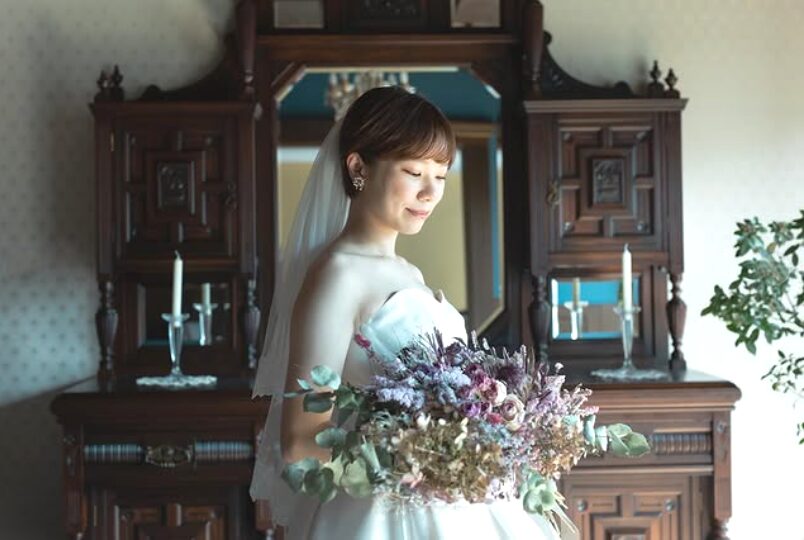 ウェディングドレスを纏い、ドライフラワーの大きなブーケを手にしたショートヘアの花嫁。クラシックな木製家具の前での室内ポートレート。