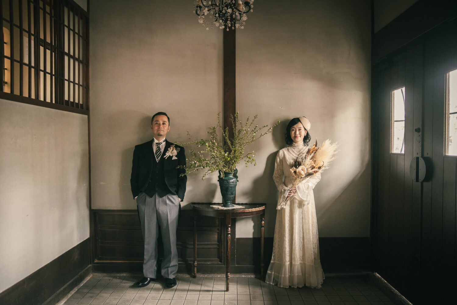 町家スタジオの室内で、窓からの光を受けて並ぶタキシードの新郎とヴィンテージドレスの新婦