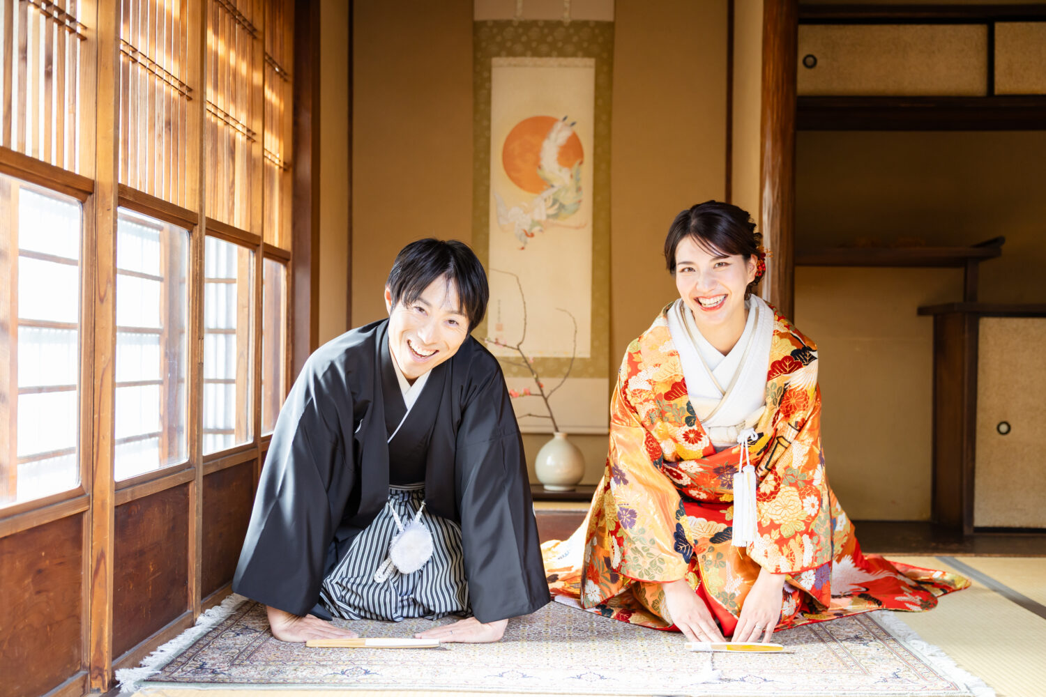 京町家の座敷で扇子を前に置き、三つ指をついて挨拶する色打掛と紋付羽織袴の新郎新婦