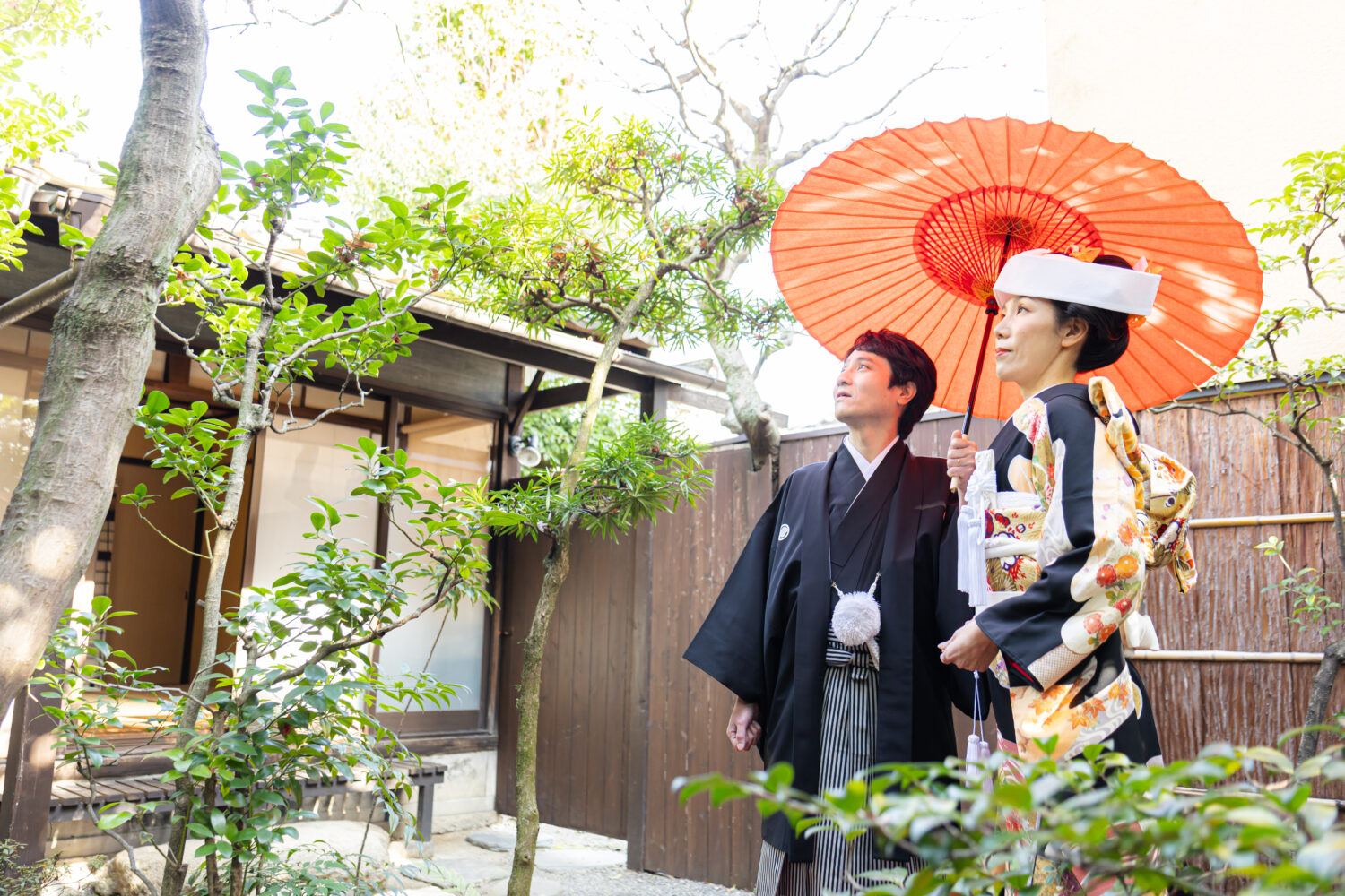 京町家の坪庭で朱色の和傘をさし、黒引き振袖と紋付羽織袴で寄り添う新郎新婦