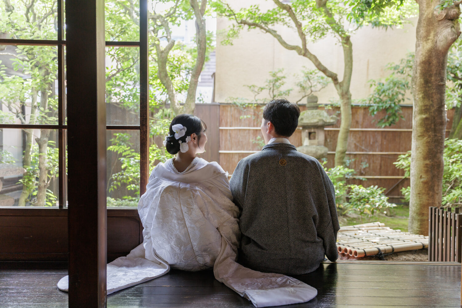 京町家の縁側に並んで座り、坪庭の新緑を眺める白無垢と紋付羽織姿の新郎新婦の後ろ姿