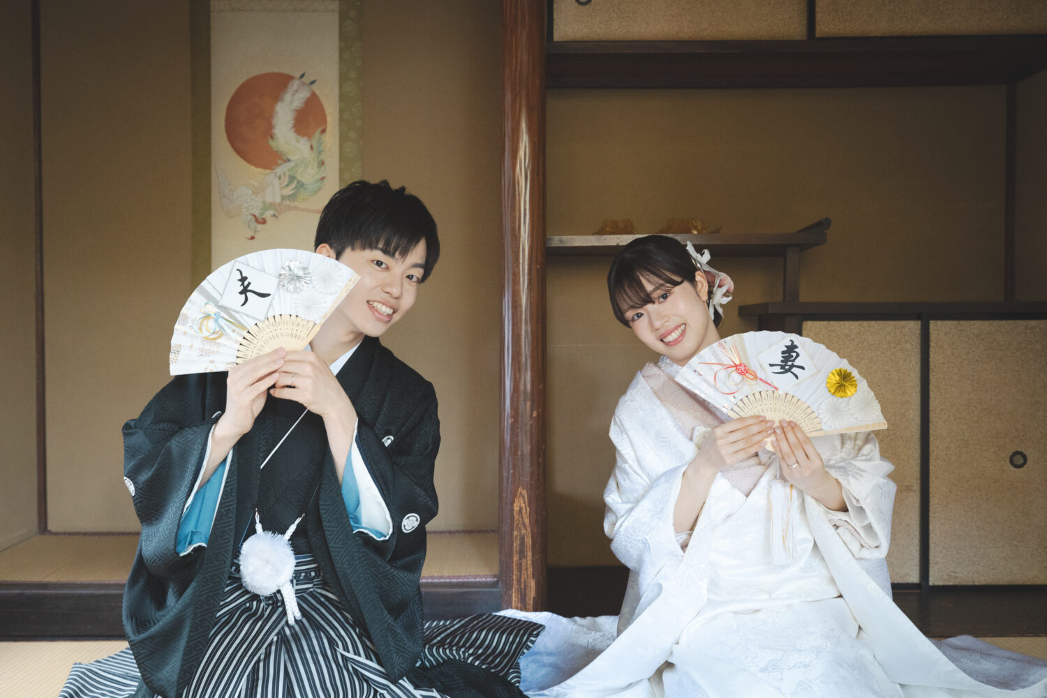 京町家の座敷で「夫」「妻」と書かれた扇子を手に笑顔を見せる白無垢と紋付羽織袴の新郎新婦
