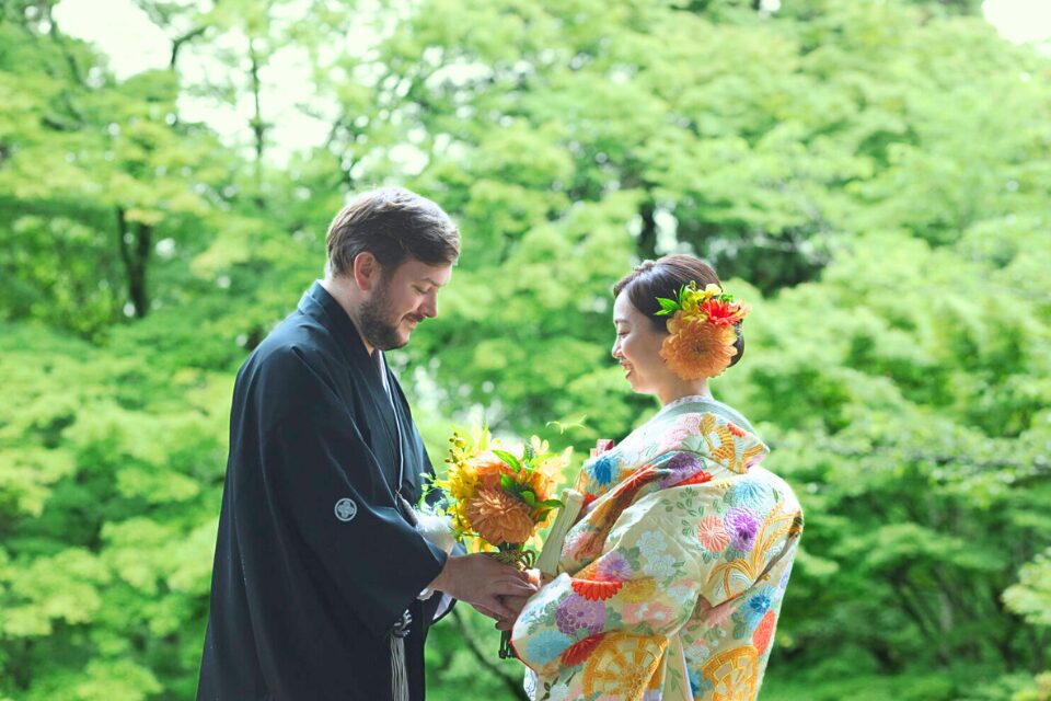 毘沙門堂の新緑の木々を背に、オレンジのブーケを持つ淡い色の色打掛の花嫁と、その手を取る紋付袴の新郎