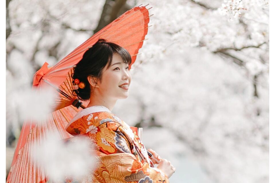 手前に桜をぼかして写した、朱色の和傘を差す金と朱の色打掛姿の花嫁