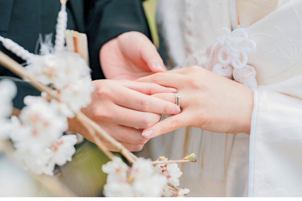 桜の枝越しに写した、白無垢の花嫁の指に指輪をはめる新郎の手元