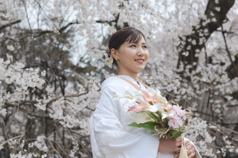 満開の桜を背に、白とピンクのカラーに桜を添えたブーケを持つ白無垢姿の花嫁